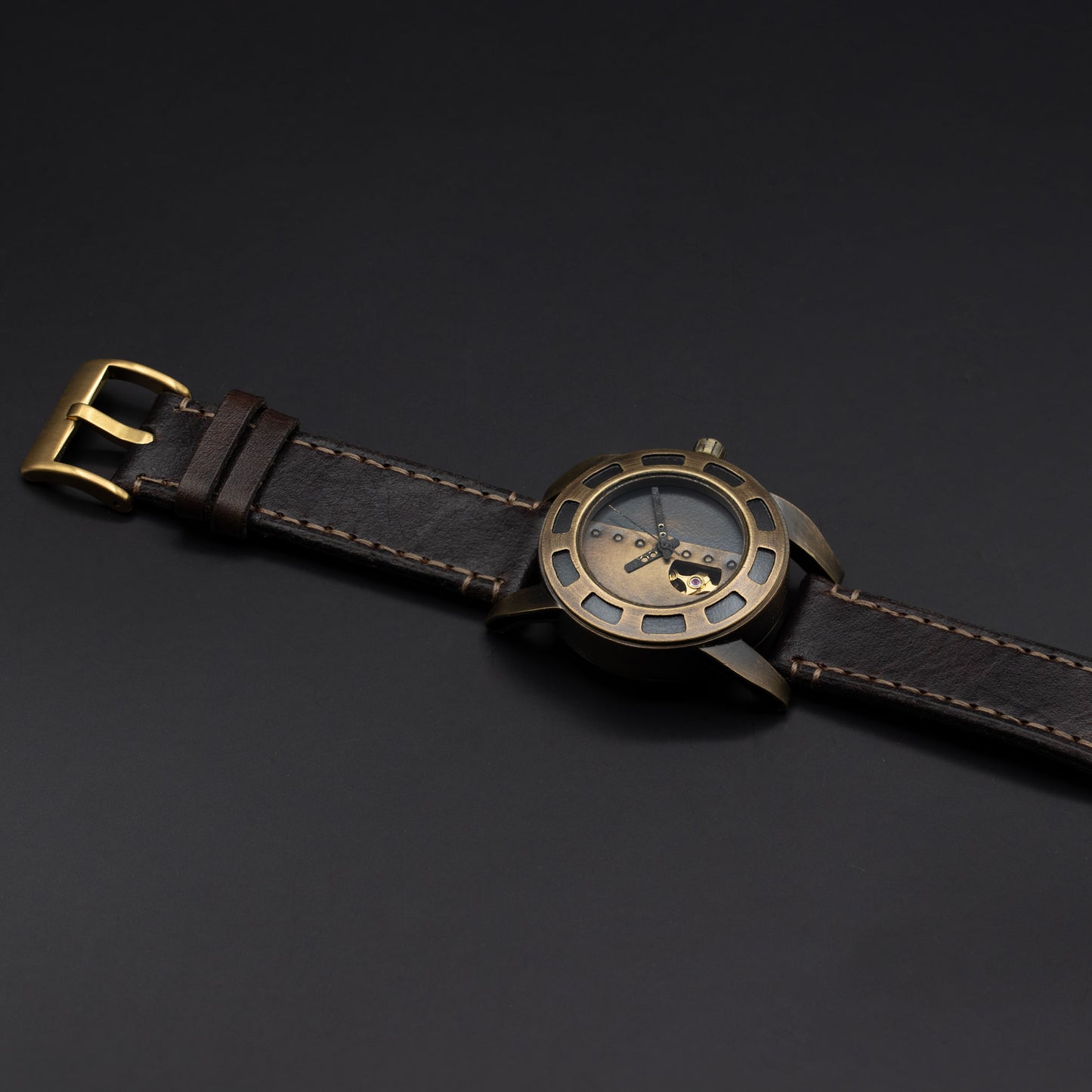 A wristwatch with a dark brown leather strap, a bronze-toned metal case, and a skeleton dial showing the inner movement, photographed on a dark gray background.