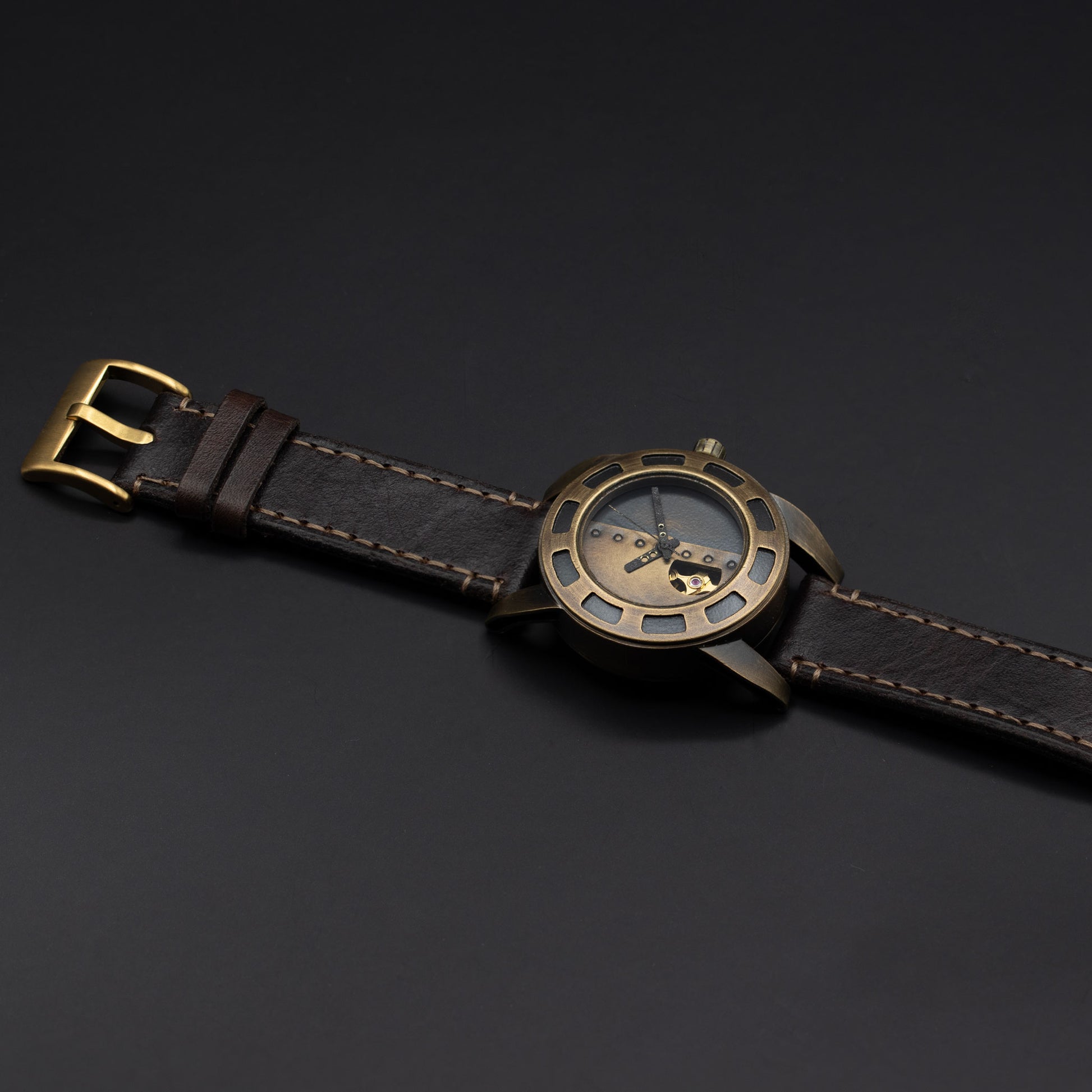 A wristwatch with a dark brown leather strap, a bronze-toned metal case, and a skeleton dial showing the inner movement, photographed on a dark gray background.