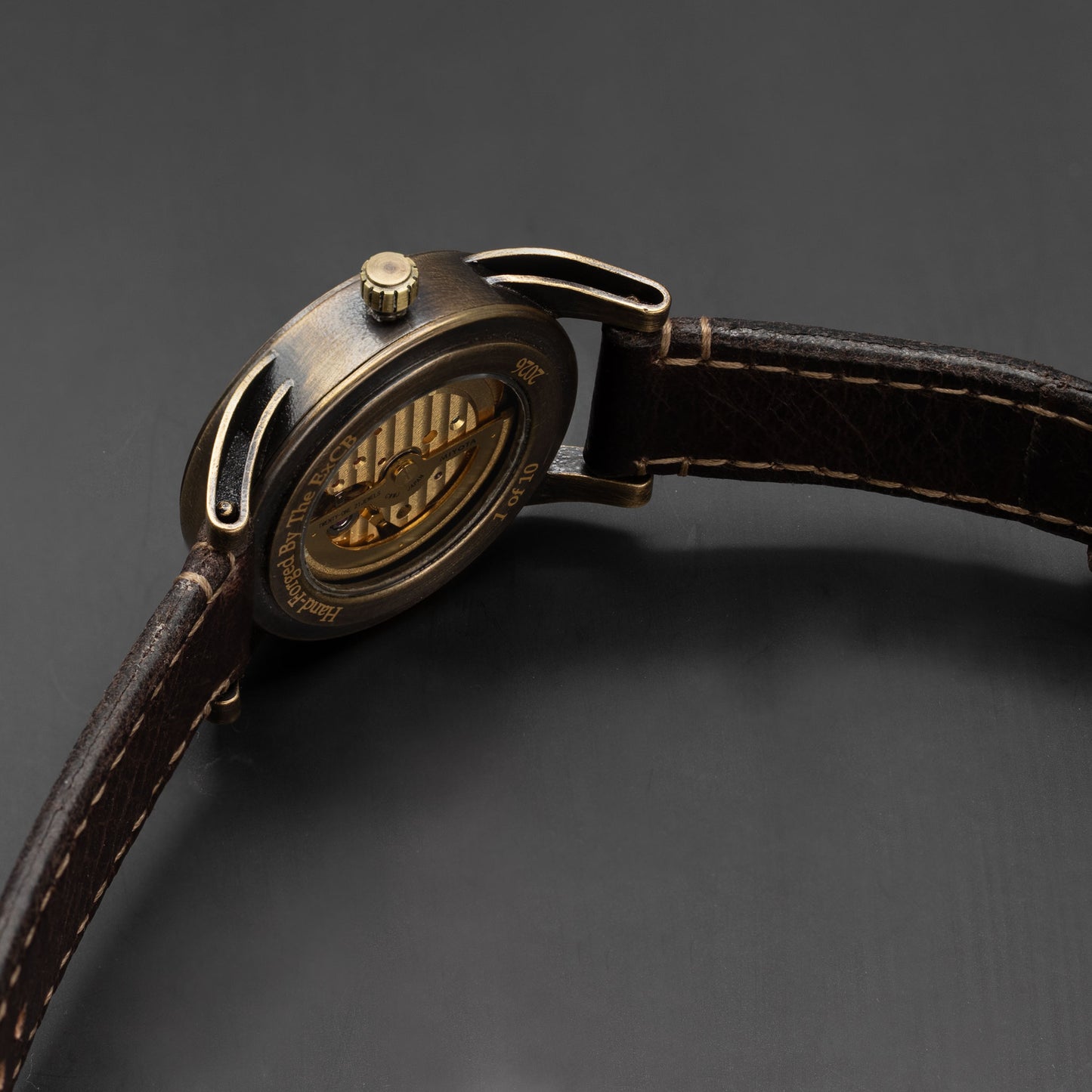Close-up of a wristwatch back showing a bronze-toned case with a visible mechanical movement and a dark brown leather strap on a dark gray background.