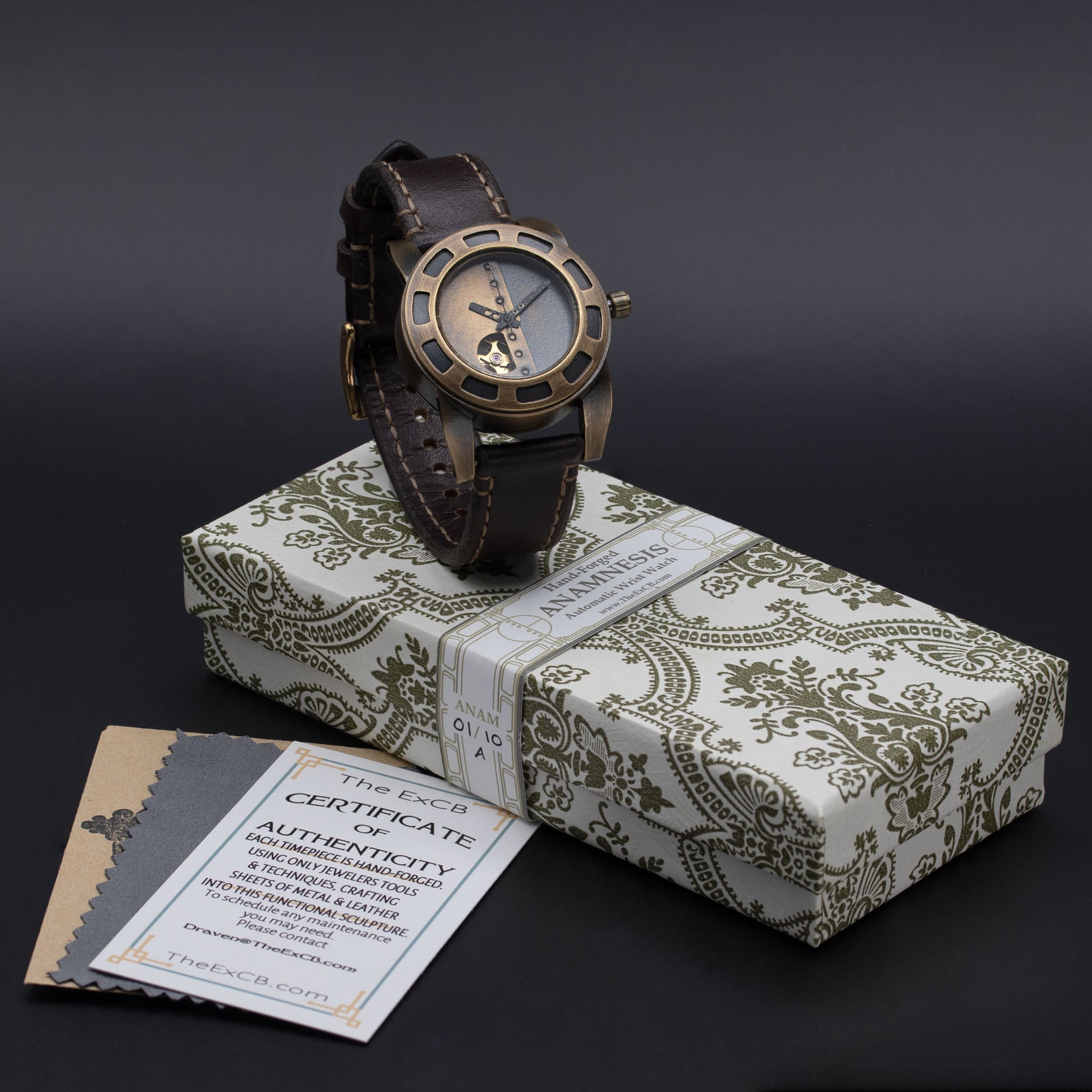 A wristwatch with a dark brown leather strap and a bronze-toned round case, displayed on top of a patterned gift box. A small authenticity card from The ExCB Collection is visible in the lower left corner against a dark gray background.