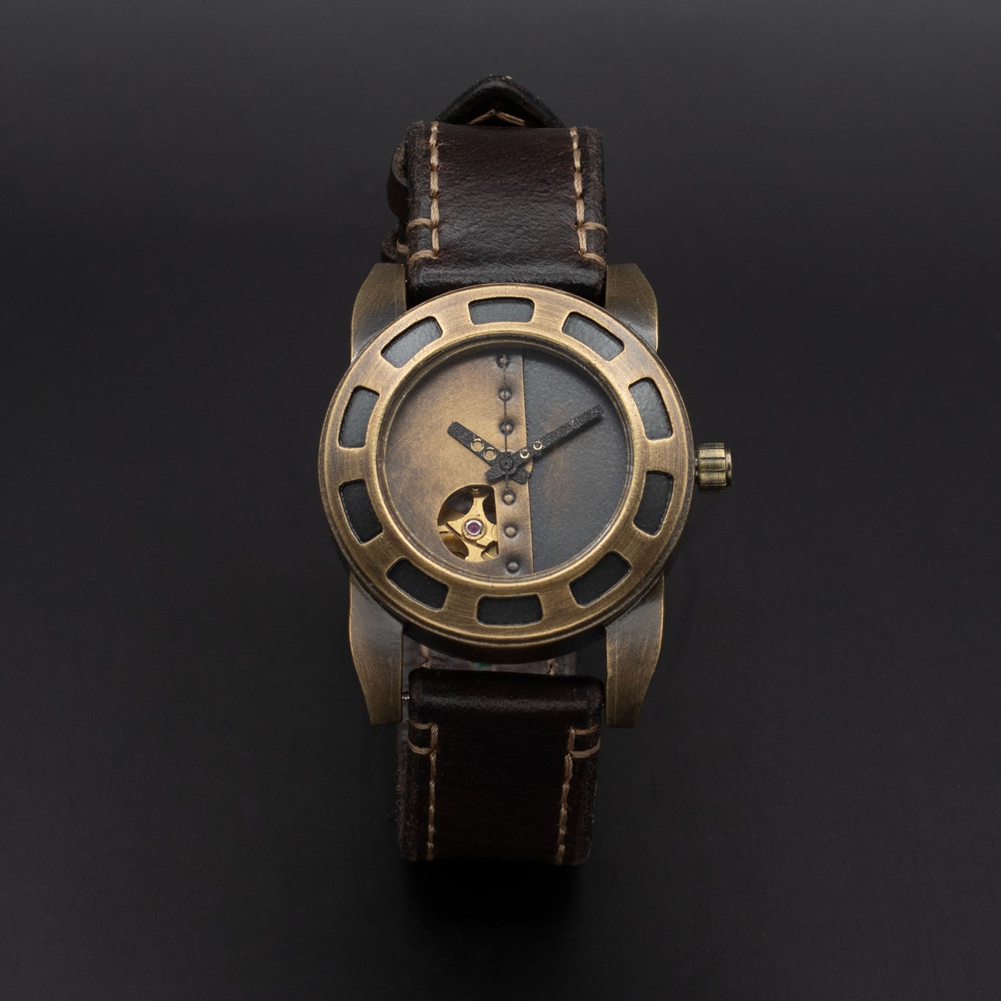 A vintage-style wristwatch with a bronze-toned metal case, skeleton dial showing a gold mechanical movement, and a dark brown leather strap with light stitching, set against a dark background.