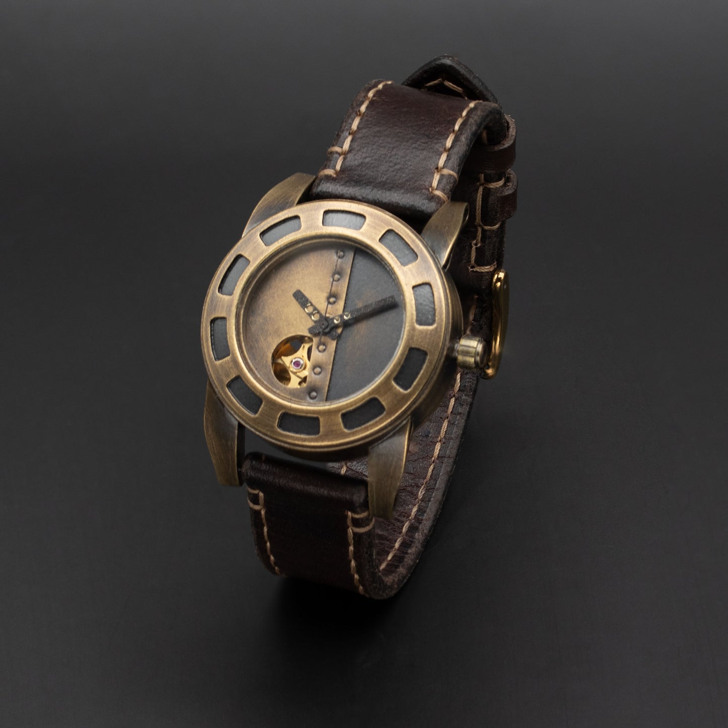 A wristwatch with a bronze-toned metal case, a skeleton dial showing a gold-toned inner mechanism, and a dark brown leather strap with contrast stitching, photographed on a dark gray background.