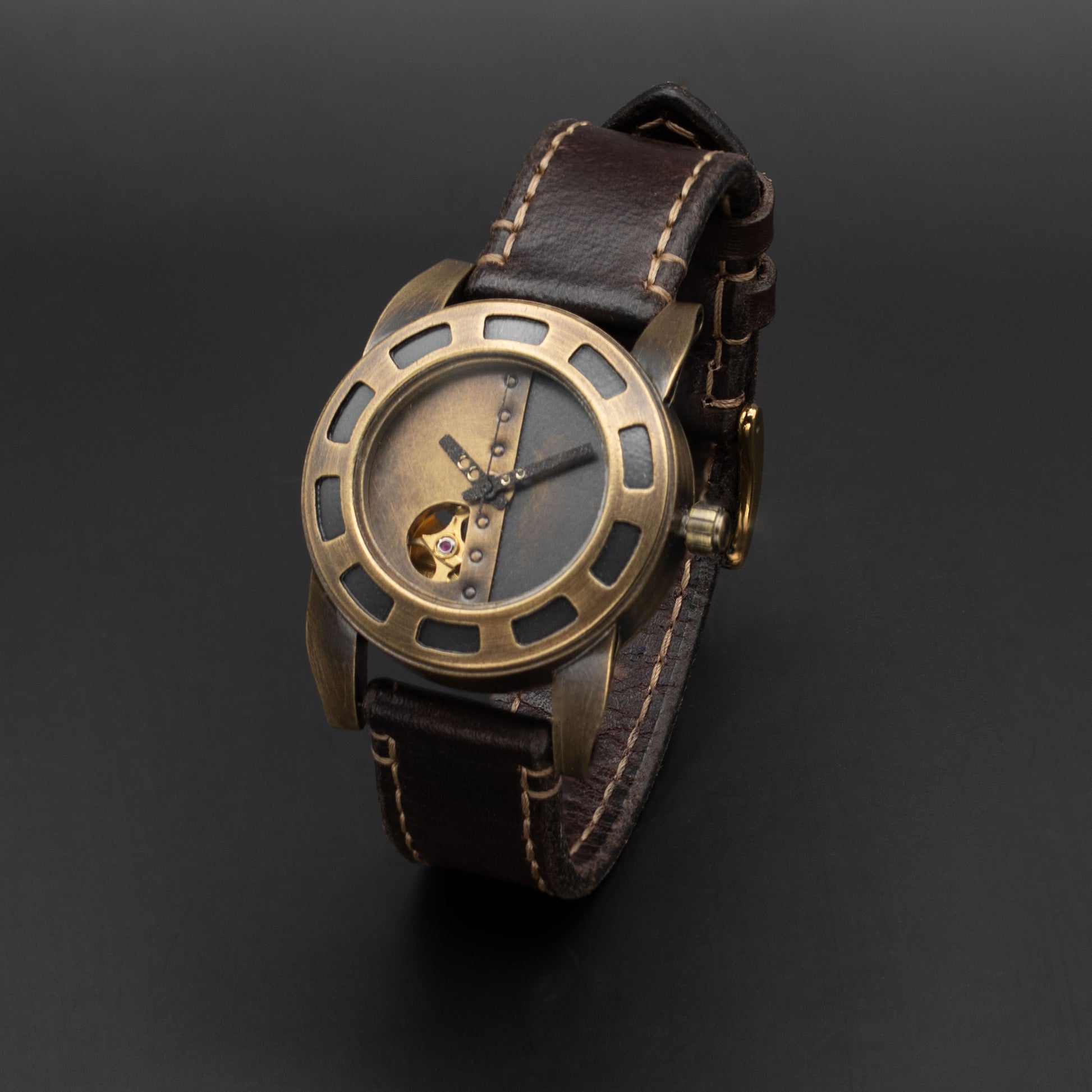 A wristwatch with a bronze-toned metal case, a skeleton dial showing a gold-toned inner mechanism, and a dark brown leather strap with contrast stitching, photographed on a dark gray background.
