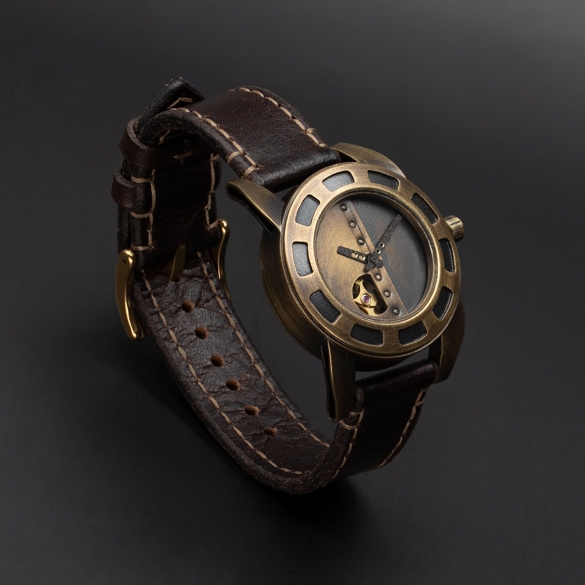 A vintage-style wristwatch with a dark brown leather strap, a bronze-toned metal case, and a skeleton dial, photographed on a dark gray background.