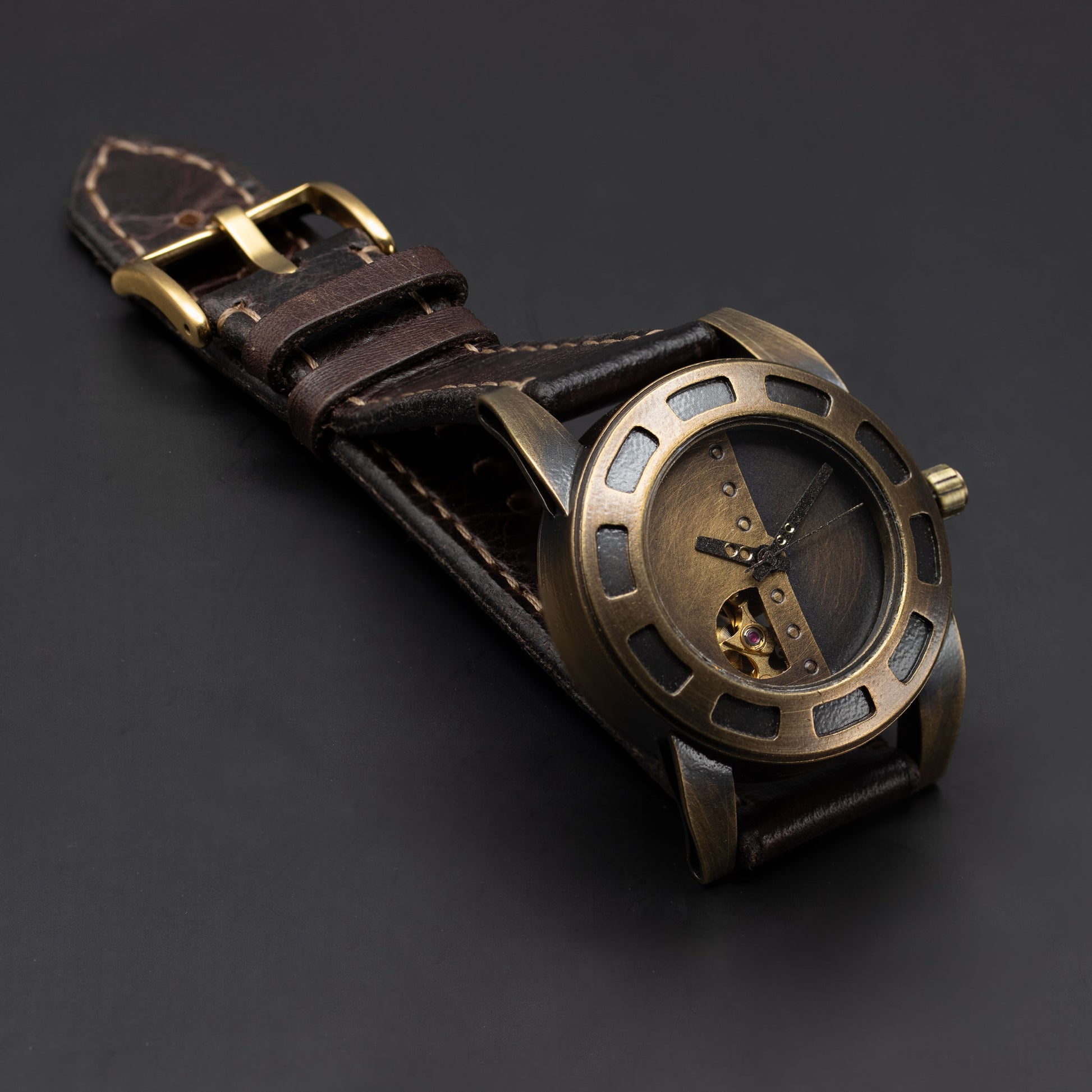 A steampunk-style wristwatch with an antique bronze-toned metal case, skeleton dial, and a dark brown leather strap, displayed on a dark gray background.